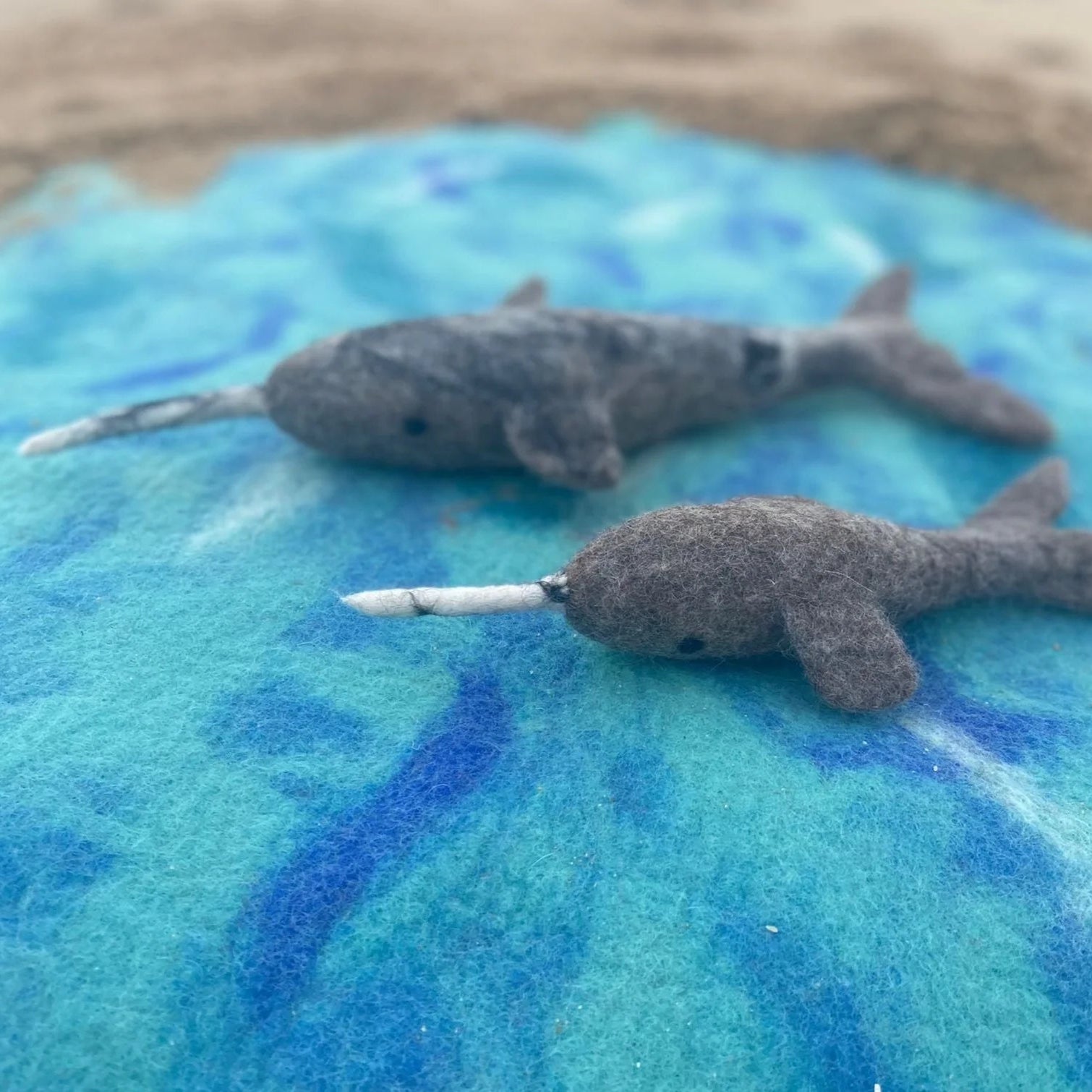 Two small gray felt narwhals with white horn-like spikes resting on a blue and turquoise fabric surface, with a blurred sandy background.