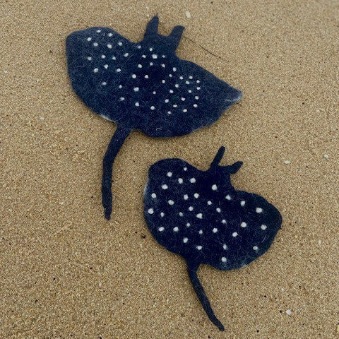 Two black stingray-shaped felt accessories with white spots, placed on a sandy beach background.