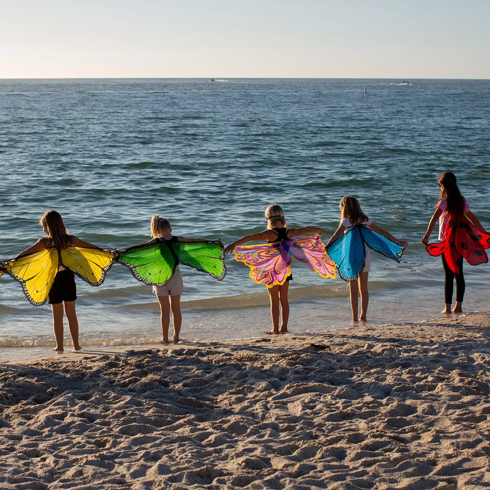 Five children with colorful costume wings standing on a sandy beach with ocean in the background