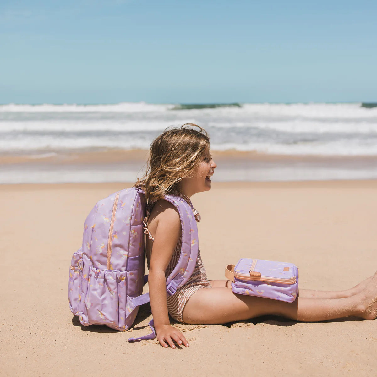 on the beach wearing Fox & Fallow backpack for kids, perfect for school, daycare and everyday use. Spacious, practical and designed for little adventures. Personalisation of embroidery with name also available on bags in 2 placements. This print is unicorns with matching lunch bag