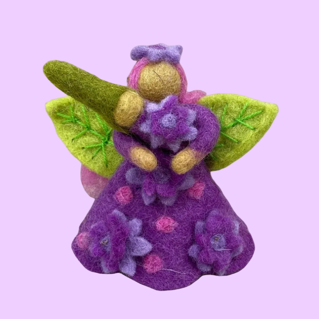 A handcrafted felted wool fairy figurine holding a purple flower bouquet. The fairy features a purple dress with pink and purple floral patterns, olive green wings, and a gold-colored head.