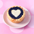 A small baked tart or pastry on a white plate, featuring a golden crust and a heart-shaped cream filling surrounded by a dark blueberry border, set against a pink bokeh background.