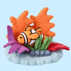 A felt fish toy with an orange body, white stripes, and black outlines, nestled among orange, purple, green, and red felt seaweed on a gray base, set against a light blue background.