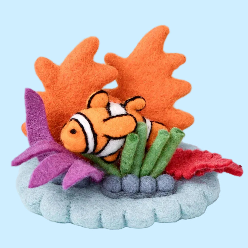 A felt fish toy with an orange body, white stripes, and black outlines, nestled among orange, purple, green, and red felt seaweed on a gray base, set against a light blue background.