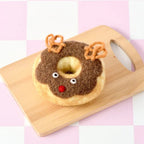 A novelty donut-shaped felt decoration shaped like a reindeer, displayed on a small wooden cutting board against a pink and white checkered background.