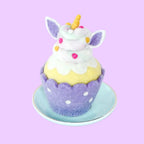 A cute unicorn-themed cupcake felt decoration sitting on a small light blue saucer. The cupcake has a purple polka-dotted wrapper, a yellow cake body, white frosting, purple unicorn ears, and a small gold horn.
