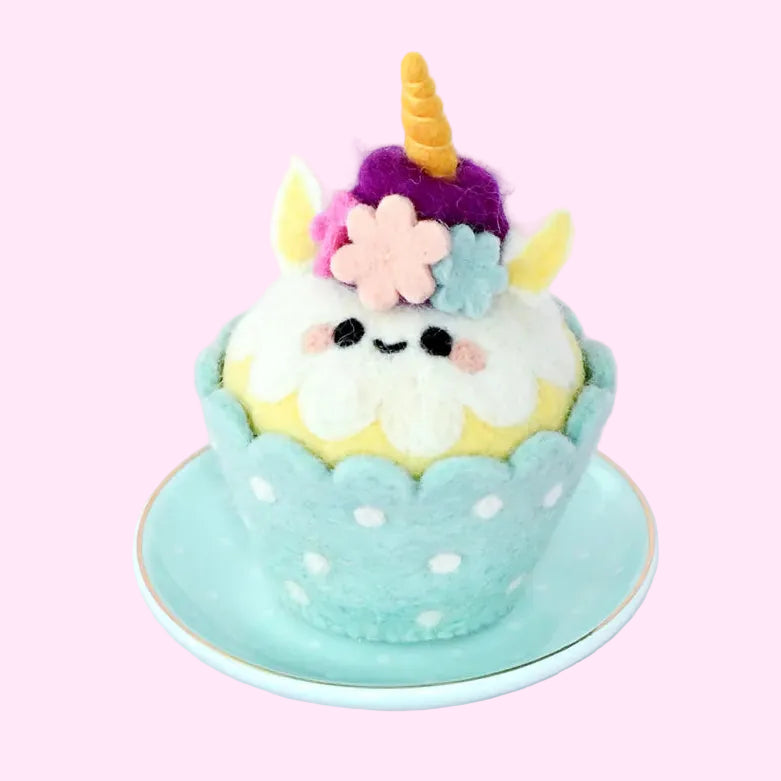 A cute, handmade felt cupcake toy with a smiling face, decorated with a purple horn, pink flower, and blue leaves, sitting on a light blue saucer against a pale lavender background.