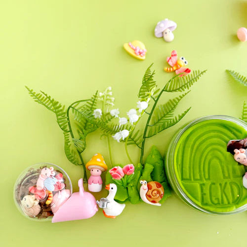 Garden sensory play accessories including green playdough, shovel, watering can, flowers and tiny garden charms arranged for tactile small world play
