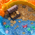 A treasure-themed sensory or play area arranged with blue and orange fabric, filled with rice, gold-colored coins, corn kernels, small stones, a toy chest, keys, starfish, and sea creature pieces.