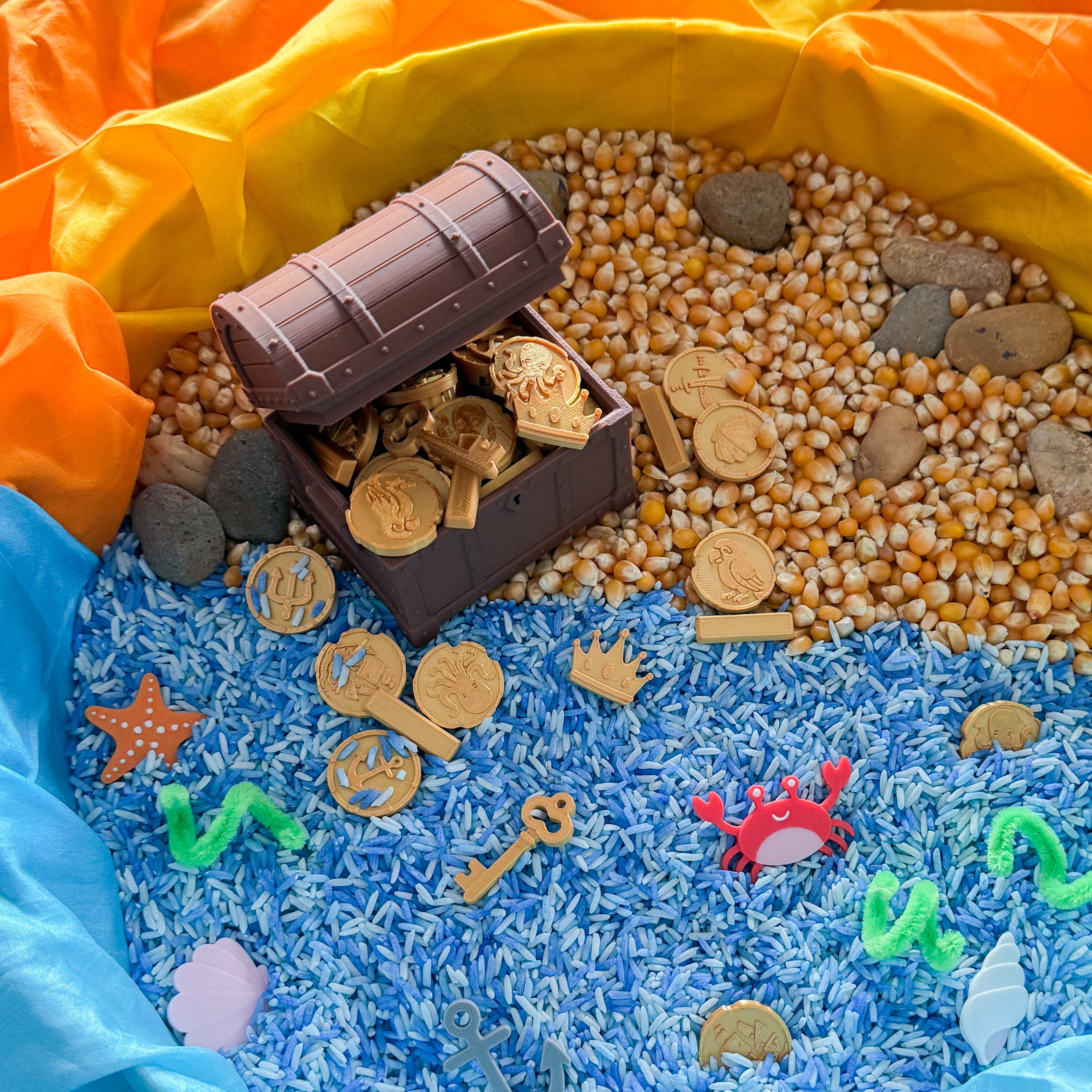 A treasure-themed sensory or play area arranged with blue and orange fabric, filled with rice, gold-colored coins, corn kernels, small stones, a toy chest, keys, starfish, and sea creature pieces.
