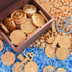 A wooden treasure chest filled with gold pirate coins, surrounded by scattered gold coins, keys, and an anchor on blue and beige rice with corn kernels.