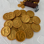 A set of gold pirate treasure coins with embossed numbers ranging from 1 to 20, displayed next to a small brown plastic treasure chest and a gray anchor on a textured white background.