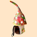 A small handmade felt mushroom house figurine with a pointed red, green, and yellow hat, white polka dots, and an open dark mouth, standing on a white textured surface against a light gray background.