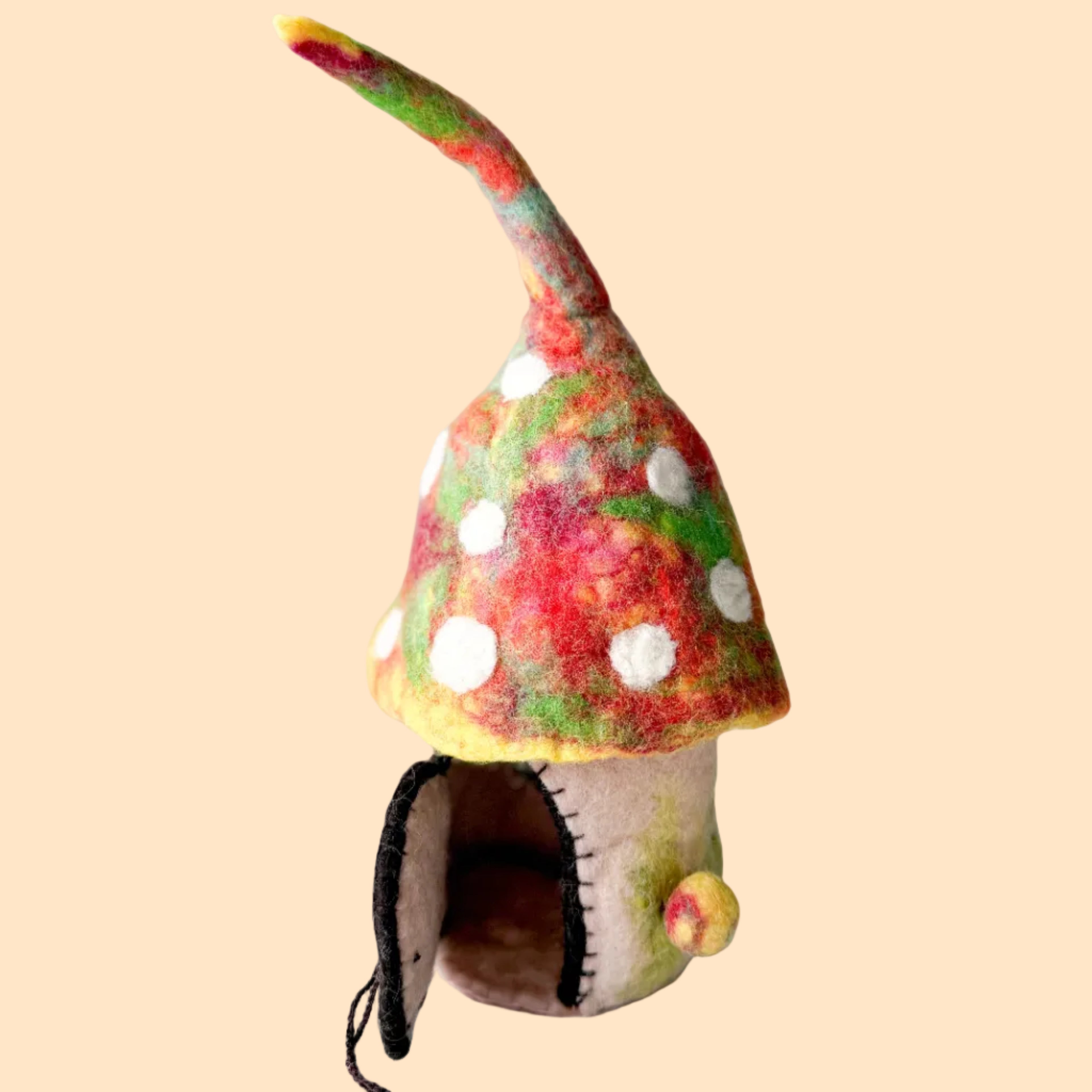 A small handmade felt mushroom house figurine with a pointed red, green, and yellow hat, white polka dots, and an open dark mouth, standing on a white textured surface against a light gray background.