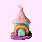 A small handmade felt house ornament with a pink pointed roof, turquoise body, rainbow arch, and purple base, photographed on a white textured surface against a light gray background.