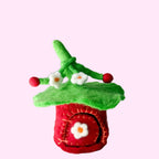 A small handmade felt house decoration with a red body, green roof, and white flower details, photographed on a white textured surface against a light gray background.
