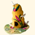 A handmade felt bee house with a yellow body, green leaves, pink flowers, and small felt bees, displayed on a textured white surface against a light gray background.