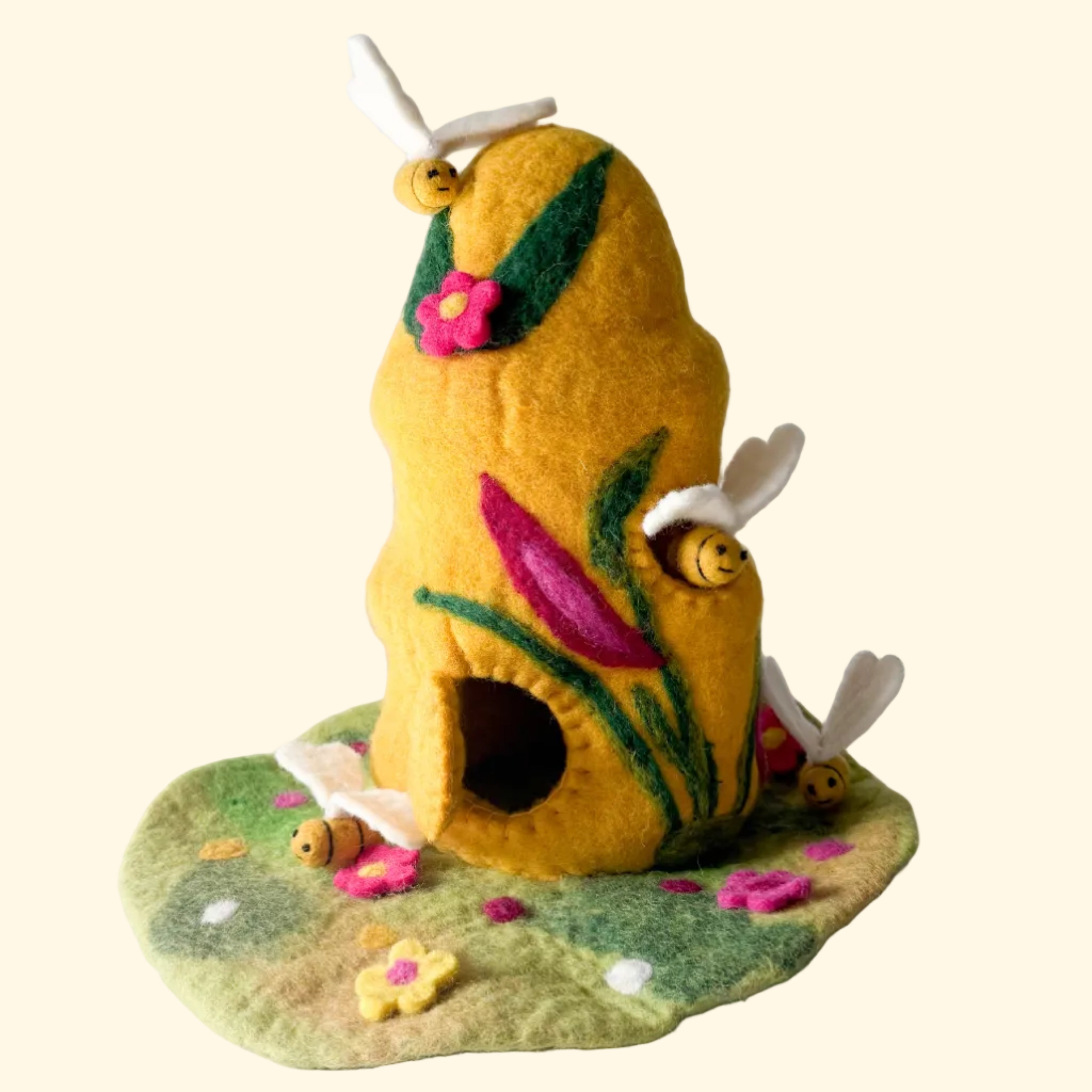 A handmade felt bee house with a yellow body, green leaves, pink flowers, and small felt bees, displayed on a textured white surface against a light gray background.
