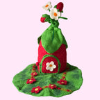 A handmade felt strawberry-shaped house decoration with a green leaf roof, red body, white flower on the door, and small white flowers on top, displayed on a textured white surface against a gray background.