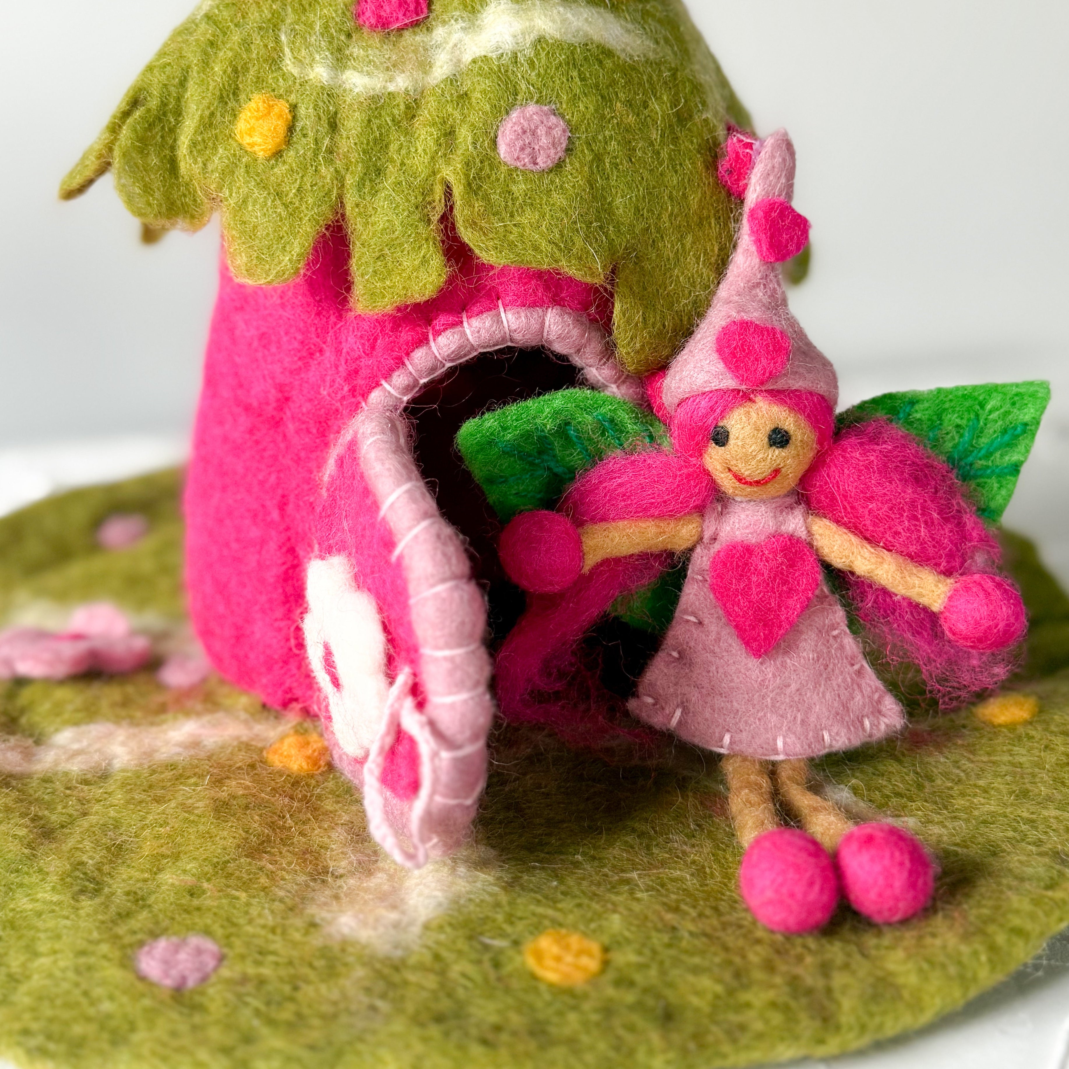 A handmade felt fairy house with a small pink fairy doll, photographed on a white textured surface against a white background.