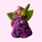 A small handcrafted felt fairy figurine in purple and pink with green wings, standing on a textured white surface against a soft light gray background.