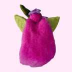 A small felted wool angel figurine with bright pink wings, olive green leaves, and a purple halo, standing on a textured white surface against a light gray background.