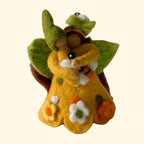 A small handmade felt creature figurine in bright yellow, decorated with green leaves, white and orange flowers, and a brown body, standing on a textured white surface against a soft light gray background.