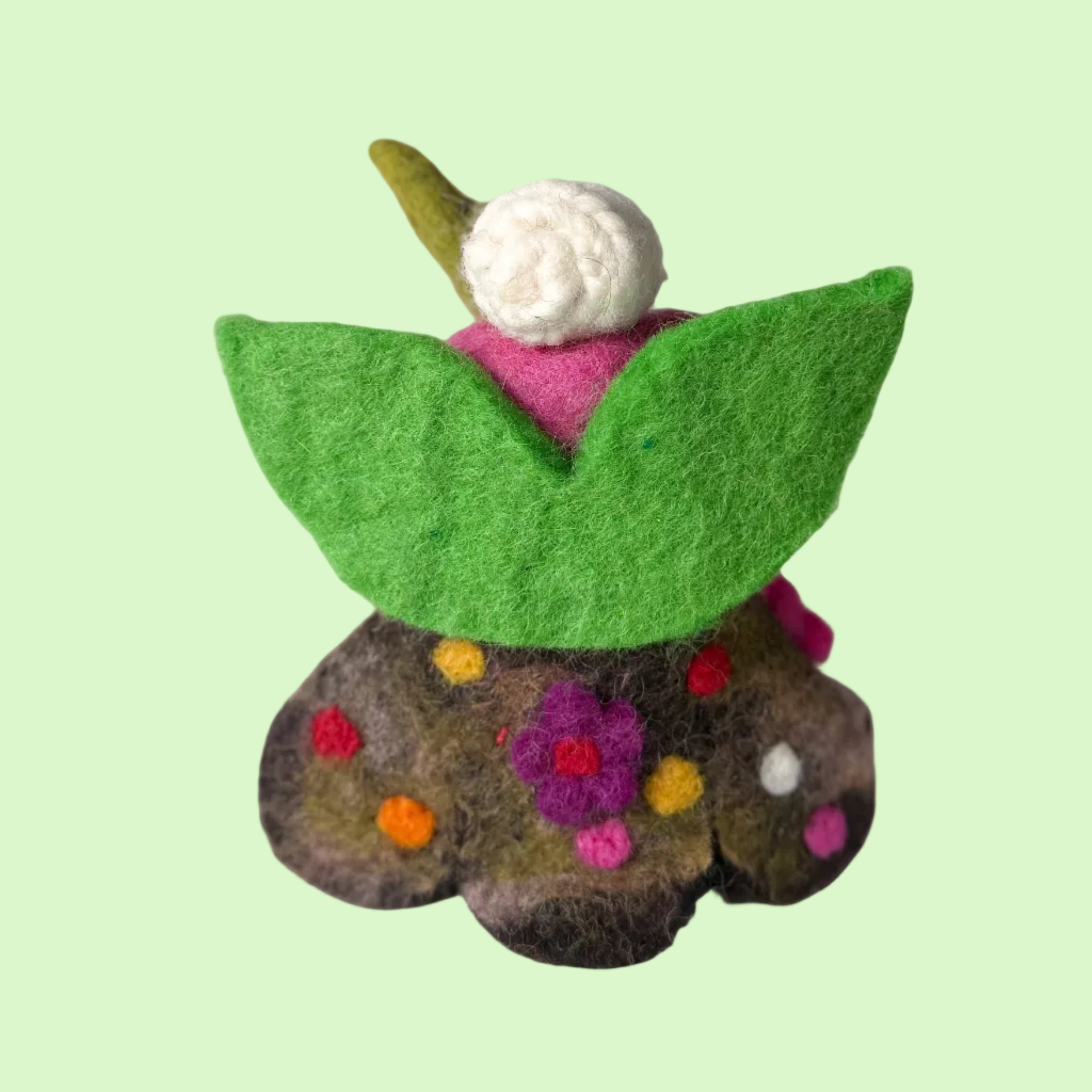 A small handmade felt flower figurine with green leaves, a pink body, a white flower, and a brown stem decorated with colorful felt dots, set against a light gray textured background.