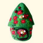 A small green felt house decoration with red flowers, ladybugs, and a bee on the front, photographed on a white textured surface against a light gray background.