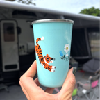 Hand holding a blue tumbler with a tiger design in front of a camper van picnic folk
