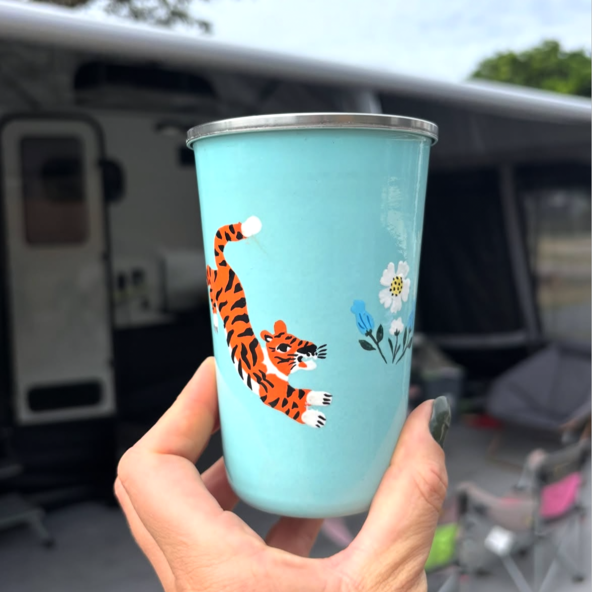 Hand holding a blue tumbler with a tiger design in front of a camper van picnic folk