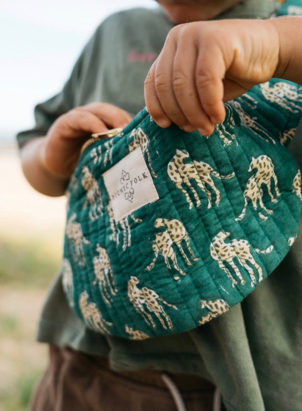 Child holding a green quilted bag with animal pattern, blurred background picnic folk crossbody bag