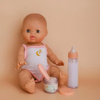 a mini coletto baby doll in a photoshoot to show off the my first tiny tummies gift pack out of the box which includes a milk bottle that disappears, and the tiny tummies jar of baby cereal with it's magic spoon. brand is tiny harlow aswell as the suit