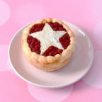 A small baked tart or pie with a golden crust, red fruit filling, and a white star-shaped topping, served on a small white plate against a pink background with soft circular bokeh.