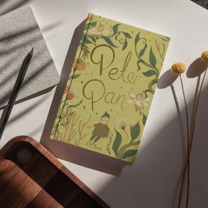photoshoot Peter Pan limited collector’s edition hardcover by J.M. Barrie with green illustrated cover, gold foil title, cloth-bound finish and decorative sprayed edges, stacked on books.