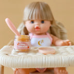 a blonde mini coletto doll sitting in rattan highchair showcasing the tiny tummies pretend play doll food jars that disappear like real food. brand is tiny harlow and tiny tummies food jar flavour is peach jelly