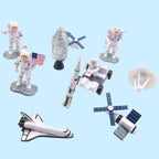 A set of small astronaut and space-themed toy figures and accessories on a plain white background, including several white astronauts, a silver rocket, a space shuttle, a satellite dish, solar panels, and a small vehicle.