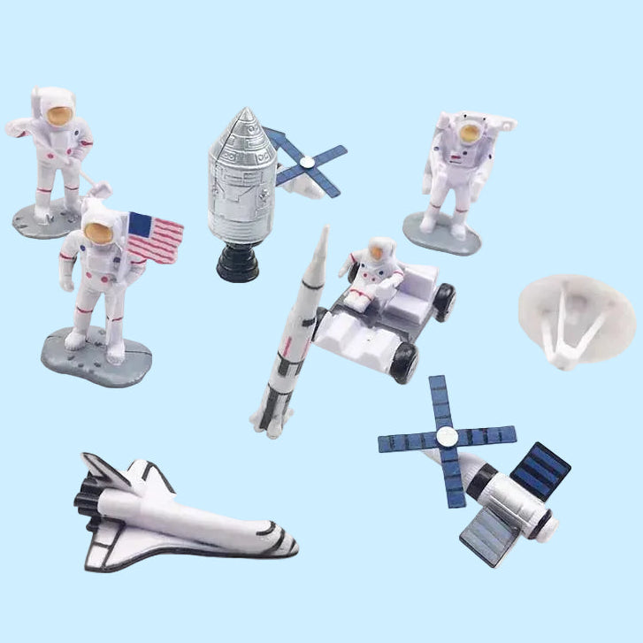 A set of small astronaut and space-themed toy figures and accessories on a plain white background, including several white astronauts, a silver rocket, a space shuttle, a satellite dish, solar panels, and a small vehicle.